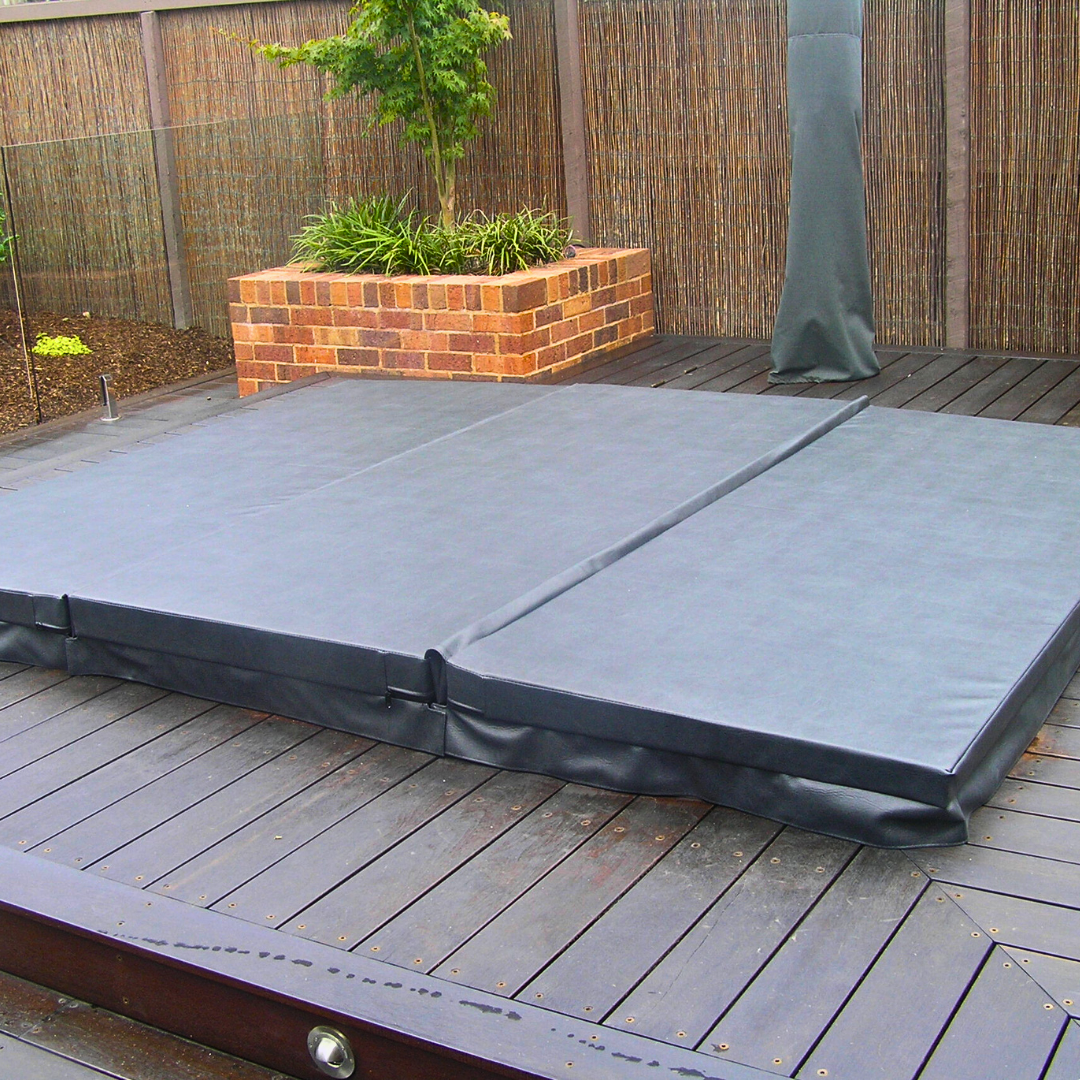A slate grey three-piece custom spa hard cover installed on a dark timber pool deck in a landscaped backyard.
