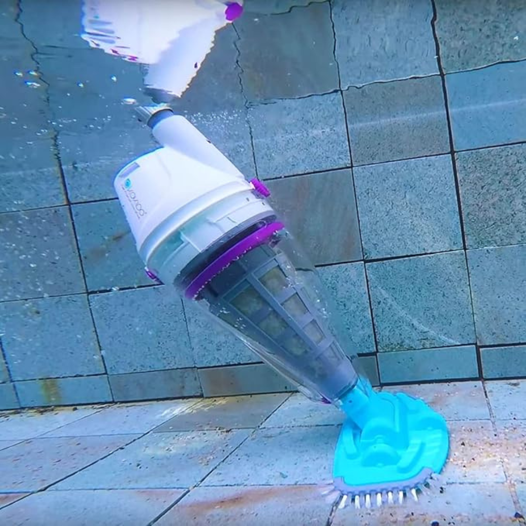 Fully submerged Kokido Telsa 50 cordless pool vacuum cleaning a grey stone tile swimming pool floor with its transparent debris canister and blue brush head visible underwater.