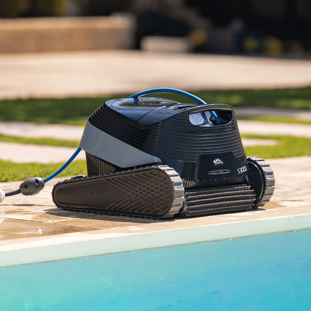 Low-angle outdoor lifestyle view of the Maytronics Dolphin S3000 robotic pool cleaner resting at the pool edge on stone paving, showcasing its front active scrubbing brush, dark navy chassis, rubber tracks, Dolphin S3000 branding, and blue power cable with anti-tangle swivel connection.