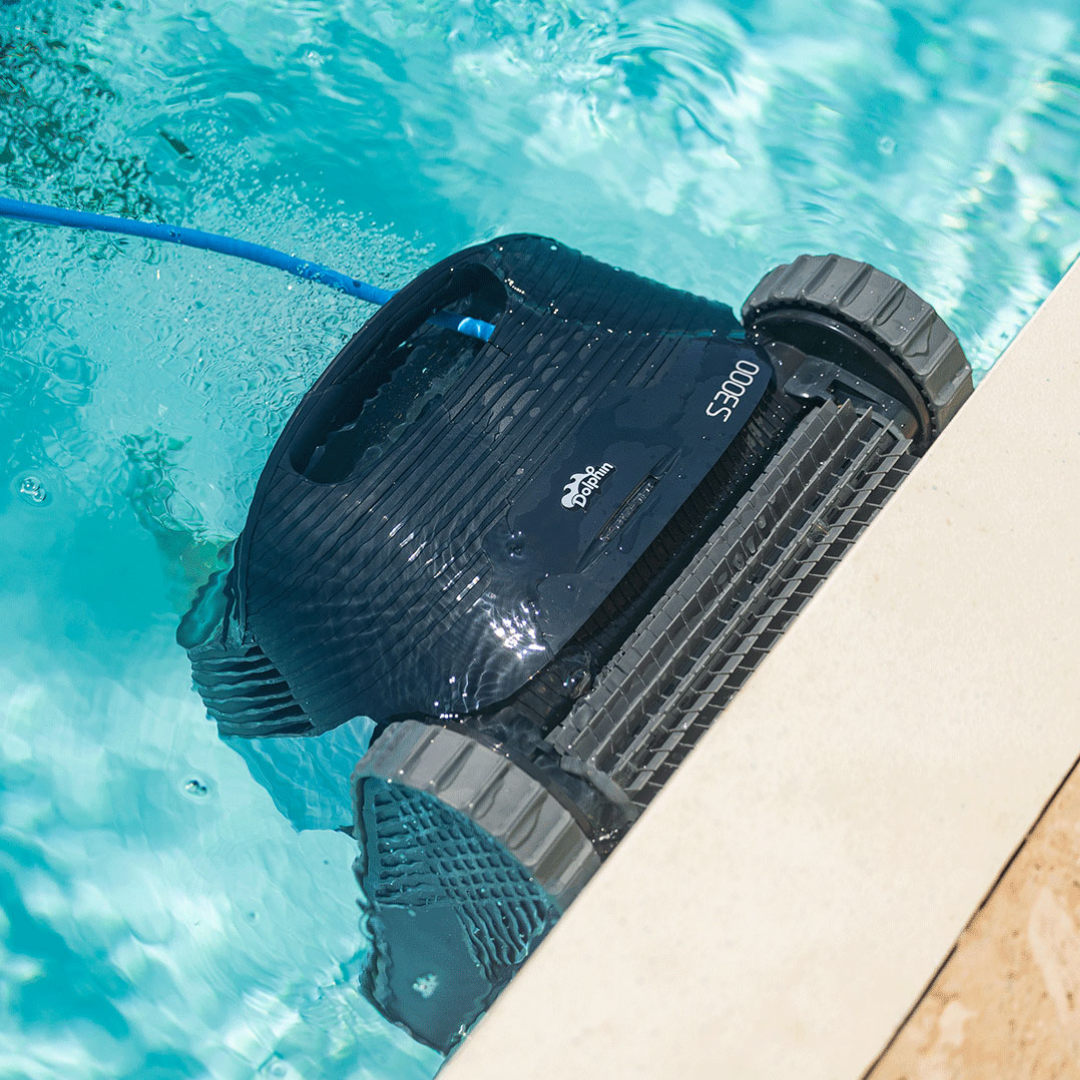Top-down outdoor lifestyle view of the Maytronics Dolphin S3000 robotic pool cleaner scrubbing along the pool waterline, showing its front active brush, dark navy chassis, S3000 branding, continuous tracks, and blue power cable floating in clear blue water.