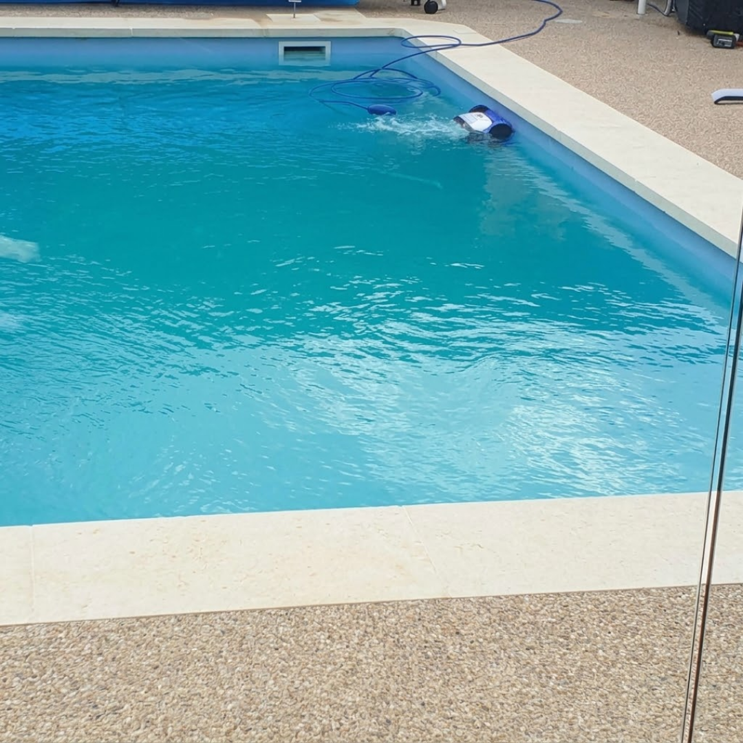Wide outdoor lifestyle view of a large residential swimming pool showing the white, grey, and blue Maytronics Dolphin S 100 robotic pool cleaner scrubbing along the waterline and pool wall.