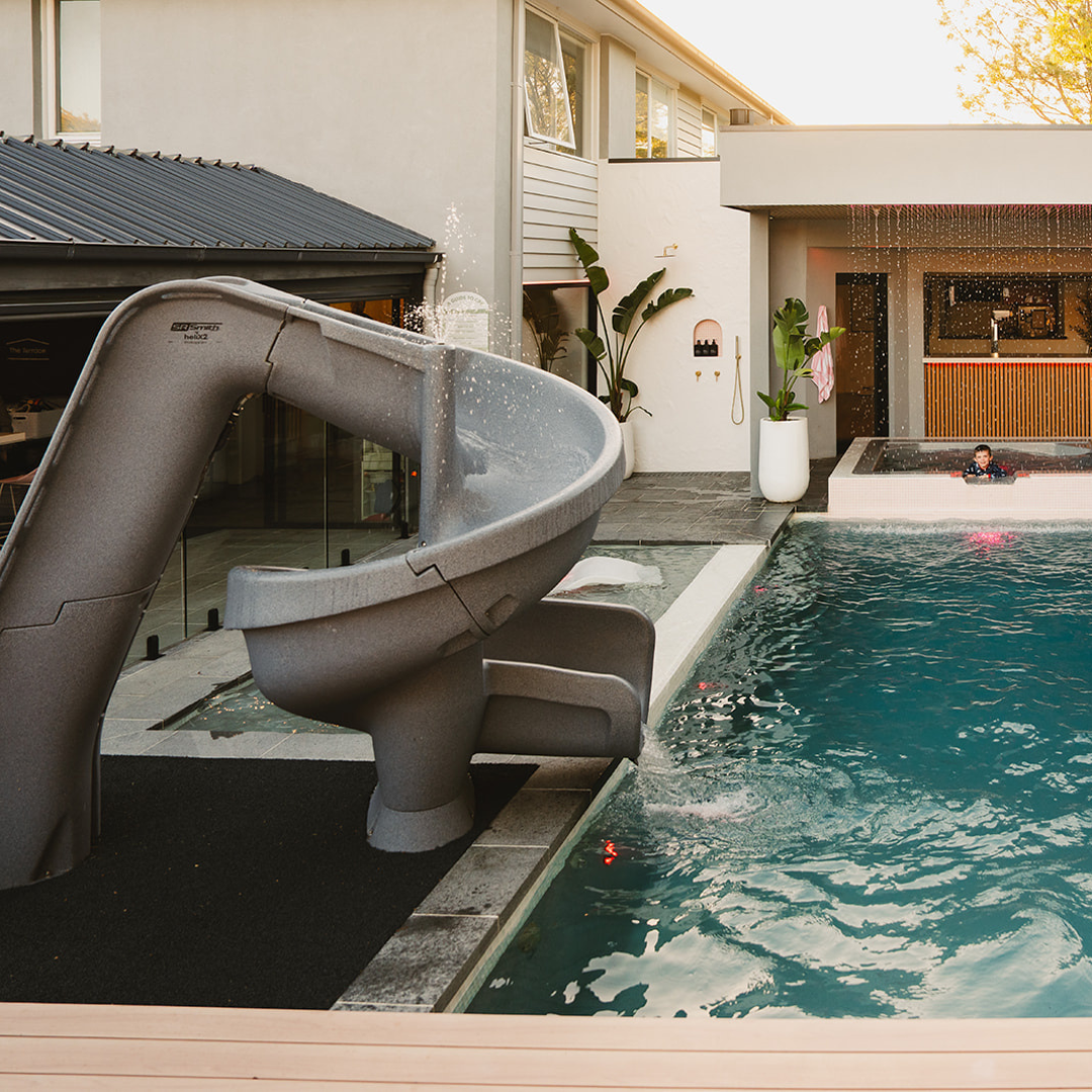 S.R.Smith Helix2 grey spiral water slide with active water spray nozzles installed next to a luxury residential inground swimming pool and outdoor cabana.
