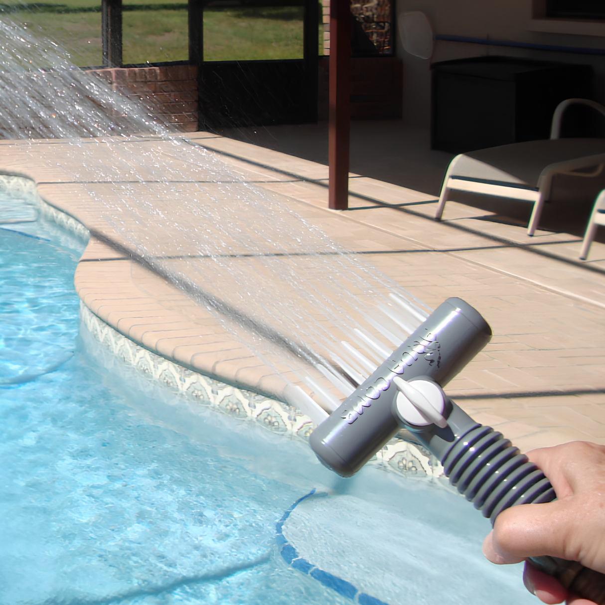 A first-person perspective shot of a hand holding a gray Aqua Comb tool, spraying a wide, flat fan of pressurized water jets across the edge of an inground swimming pool with a paved concrete deck and lounge chairs in the background.