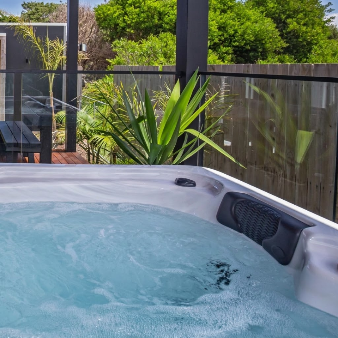 A dark charcoal black foam lounger headrest pillow installed in a bubbling, water-filled white acrylic hot tub on an outdoor deck, featuring a background with a glass safety fence, tropical plants, and a wooden boundary wall.