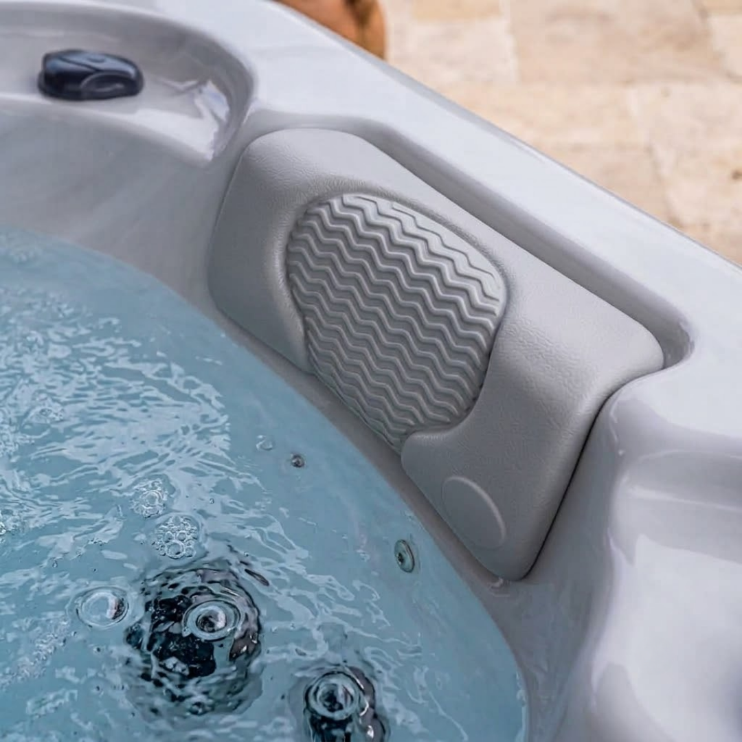 Close-up view of a light grey foam lounger headrest pillow installed in a bubbling, water-filled hot tub, showcasing its textured surface and wavy-ridge center pad alongside an outdoor stone patio area.