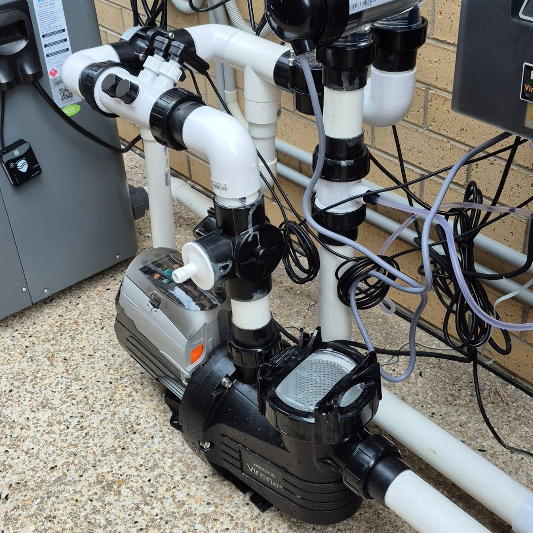 Close-up view of an outdoor pool equipment installation featuring an AstralPool Viron P320XT eVo variable speed pump connected with white PVC plumbing and valves, set against a brick wall on an aggregate concrete pad.