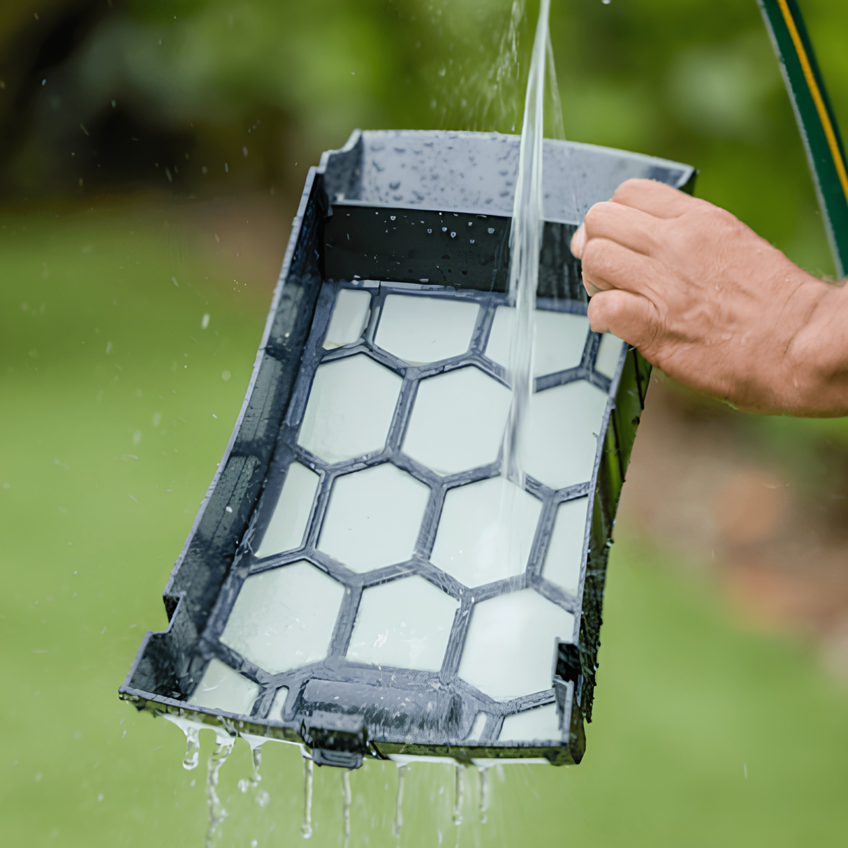 A person rinsing the black and white honeycomb filter basket tray of a Maytronics Dolphin Skimmi 300 solar pool cleaner with a garden hose.