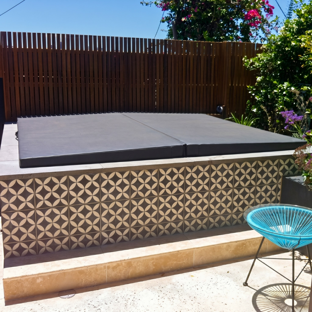 A closed dark grey custom spa hard cover installed on an outdoor hot tub clad in decorative patterned tiles.