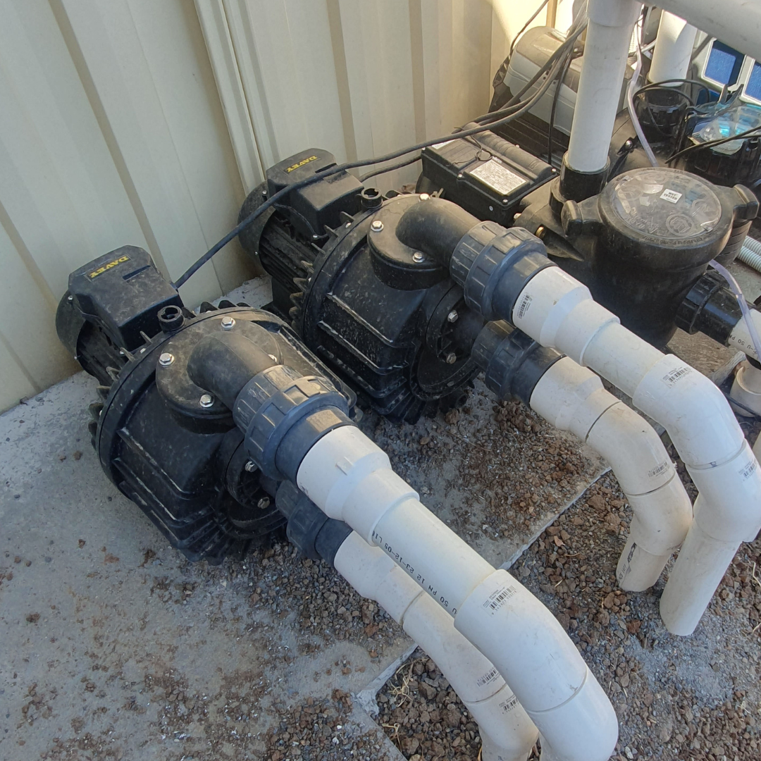 Field installation photo showing two Davey Hurricane H300M Turbo spa pumps installed side-by-side on a concrete slab against a metal wall, connected to white PVC pipework with quick-connect barrel unions and electrical conduit.
