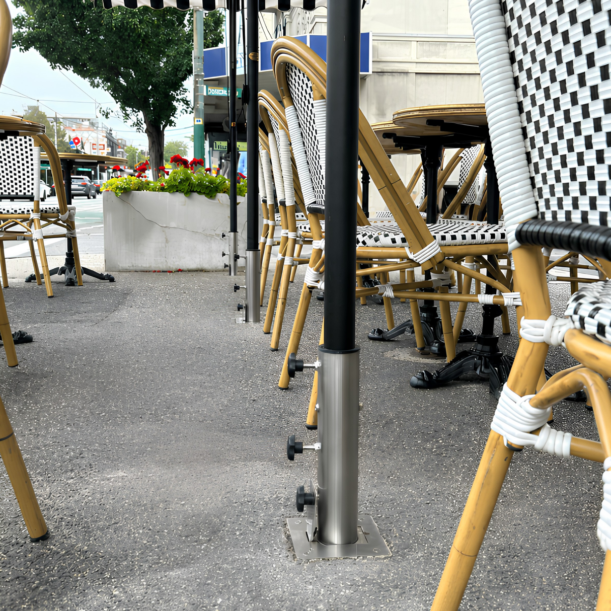 Line of heavy-duty stainless steel EzyHold commercial umbrella holder sleeves bolted flush into an asphalt sidewalk cafe patio with black bistro chairs and tables