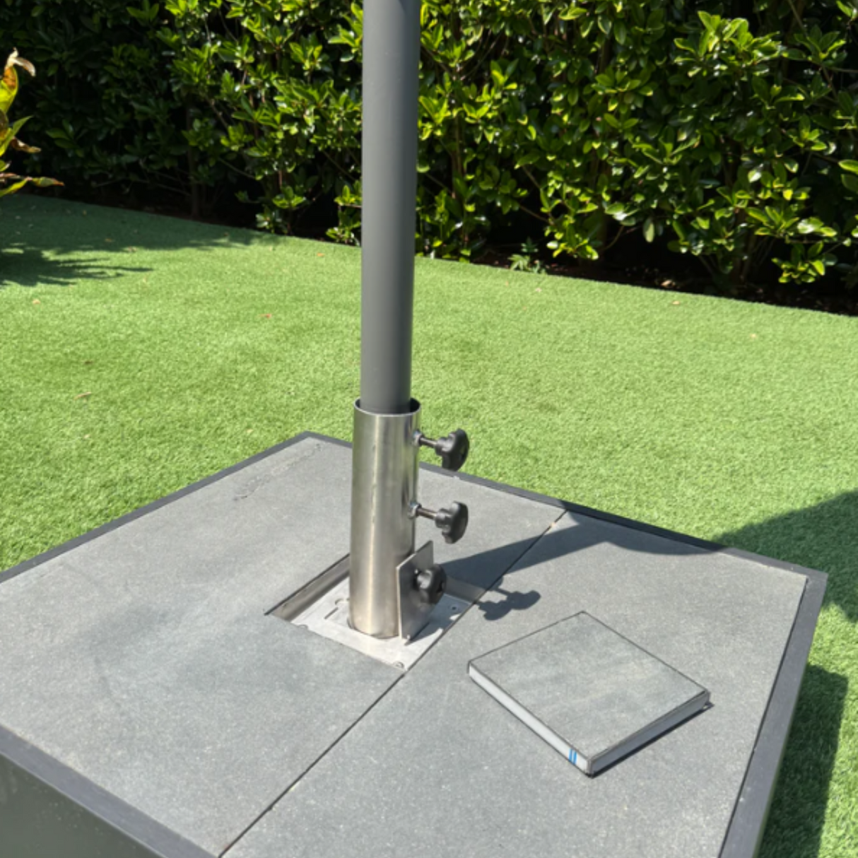 EzyHold Insert 316 marine-grade stainless steel flush-mount umbrella holder installed into stone paving tiles with the stabilizing sleeve securing a patio umbrella pole and a matching tile-filled cover plate sitting beside it.