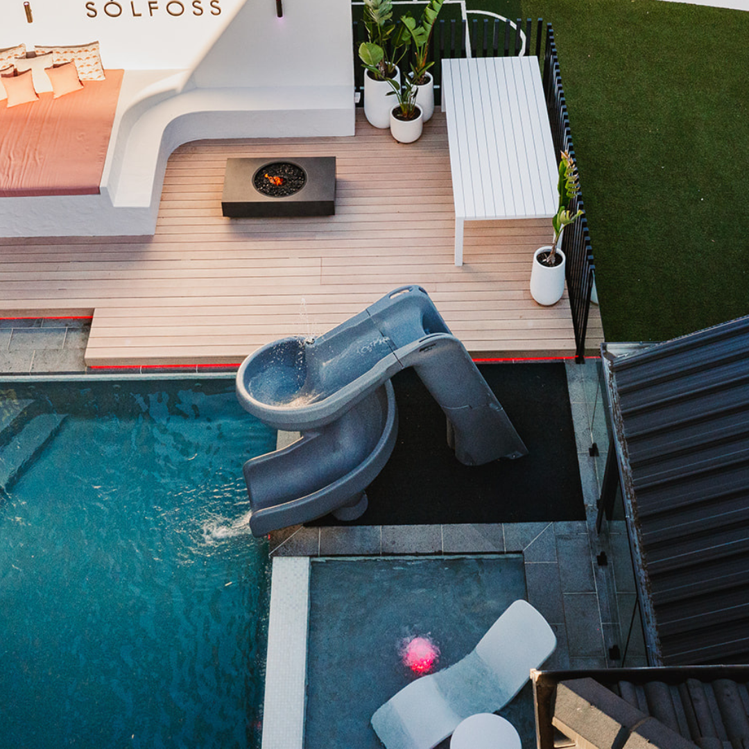 High angle view of an S.R.Smith Helix2 grey spiral water slide running into an inground swimming pool next to a modern timber deck with a built-in fire pit