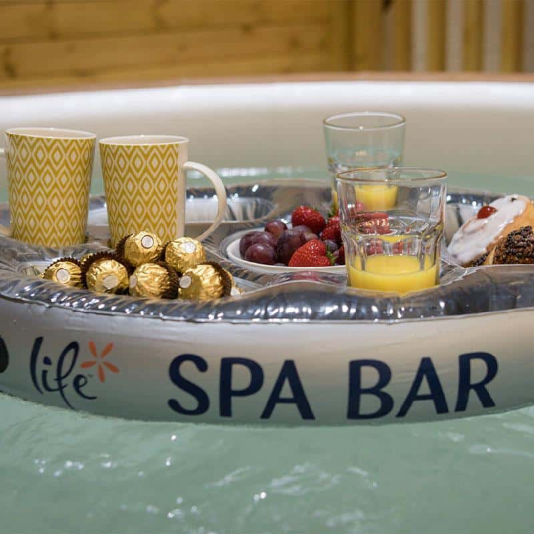 A close-up lifestyle shot of the inflatable silver Life Spa Bar floating tray drifting in hot tub water. The bar is filled with two pattern-printed coffee mugs, gold-wrapped chocolate truffles, a small plate of fresh strawberries and grapes, glasses of orange juice, and a glazed pastry.