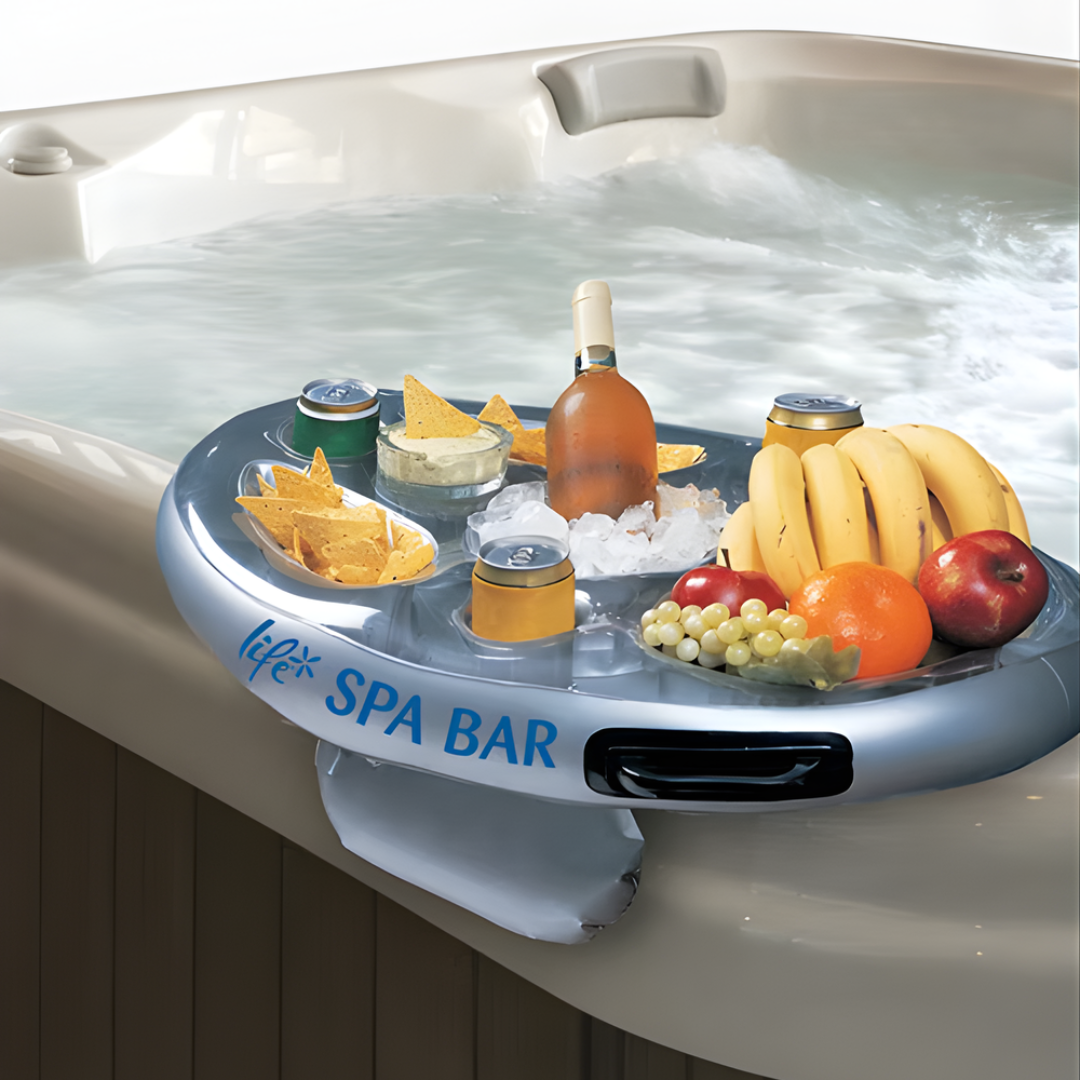 An inflatable silver Life Spa Bar snack tray and beverage holder balancing on the edge of a bubbling beige acrylic hot tub. The caddy is stocked with a wine bottle on ice, canned drinks, tortilla chips with dip, and fresh fruit, with its weighted stability pouch securely hanging over the side of the hot tub wall.