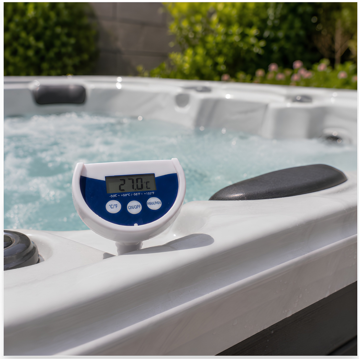 A white and blue Life Spa Essentials floating digital thermometer resting inside the shell of an active hot tub. The LCD screen displays a clear water temperature reading of 27.0°C against a background of bubbling water, dark headrests, and blurred backyard greenery.