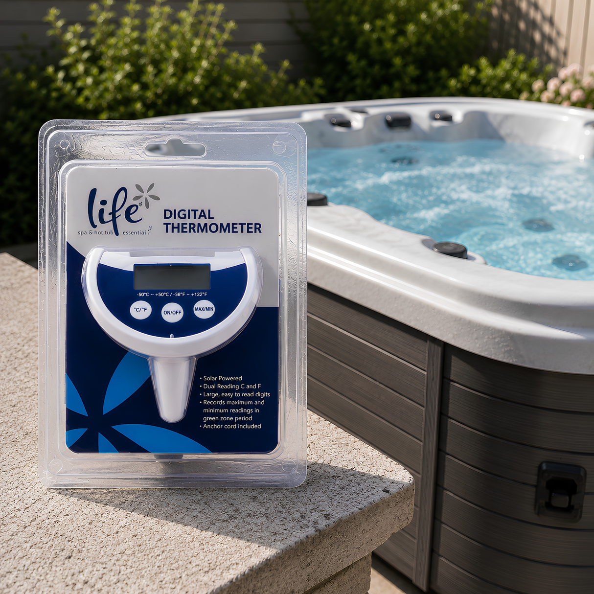 The Life Spa Essentials Floating Digital Thermometer in its clear retail clamshell packaging stands on a stone ledge next to a bubbling gray hot tub. The packaging card lists product features: Solar Powered, Dual Reading C and F, Large easy to read digits, Records maximum and minimum readings, and Anchor cord included.