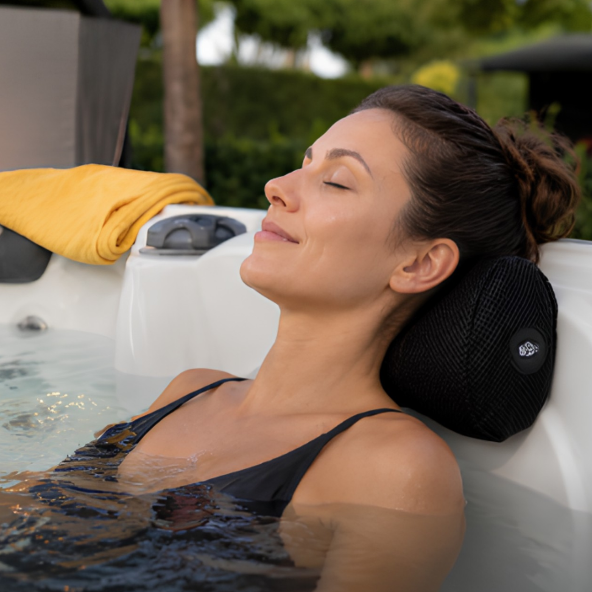 A woman in a black swimsuit relaxing with her eyes closed inside an outdoor hot tub, resting her head on a contoured black Life Spa Pillow cushion mounted to the white acrylic tub rim, with a yellow towel and blurred backyard greenery in the background.