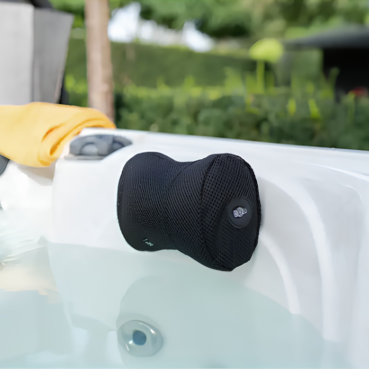 A black Life Spa Pillow installed as a neck rest on the inner white shell rim of an outdoor hot tub filled with clear water, featuring a yellow towel resting nearby on the deck and a softly blurred green garden landscaping background.