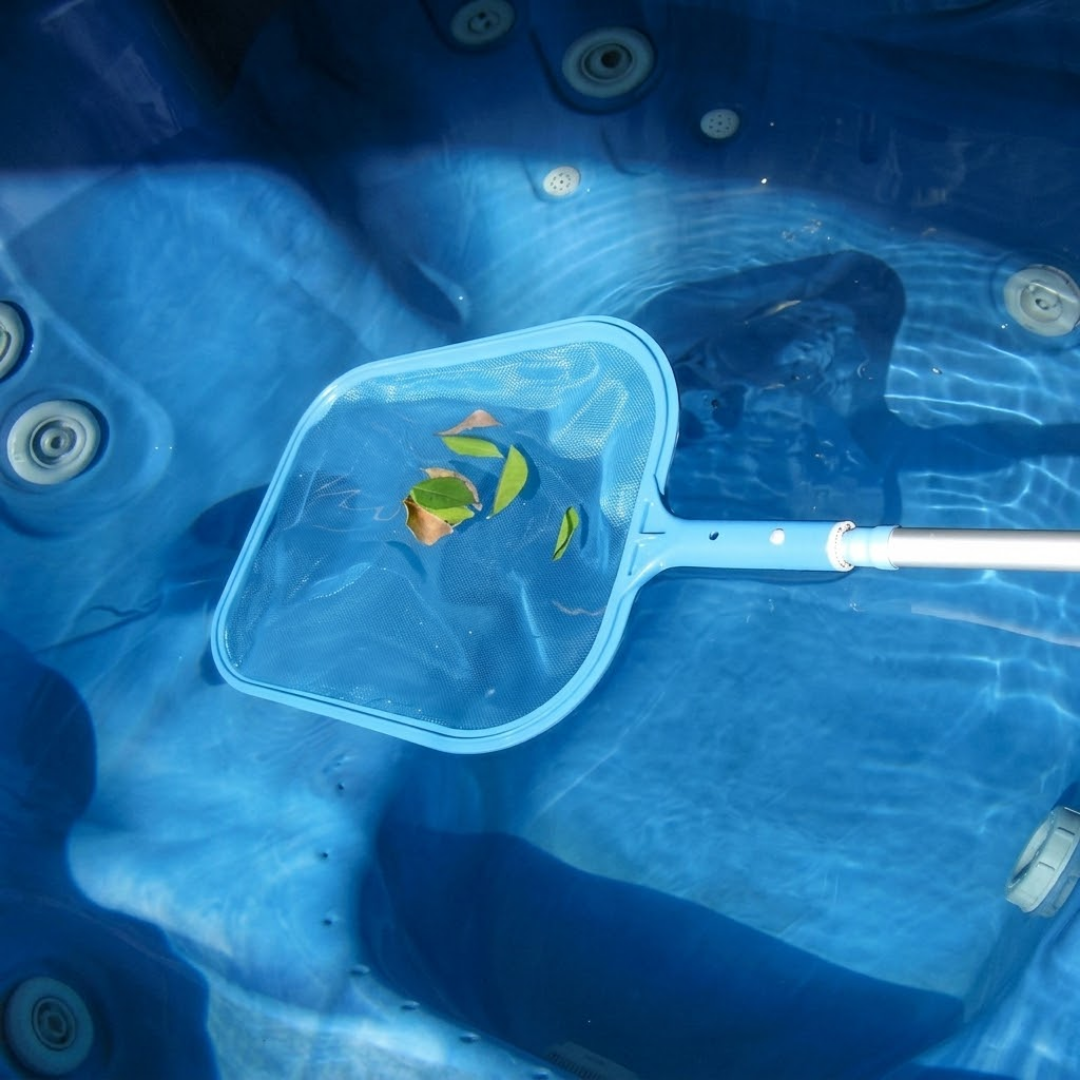 Overhead action photo of a light blue spa skimmer net attached to a telescopic pole, collecting floating leaves from the clear water inside a blue acrylic hot tub.