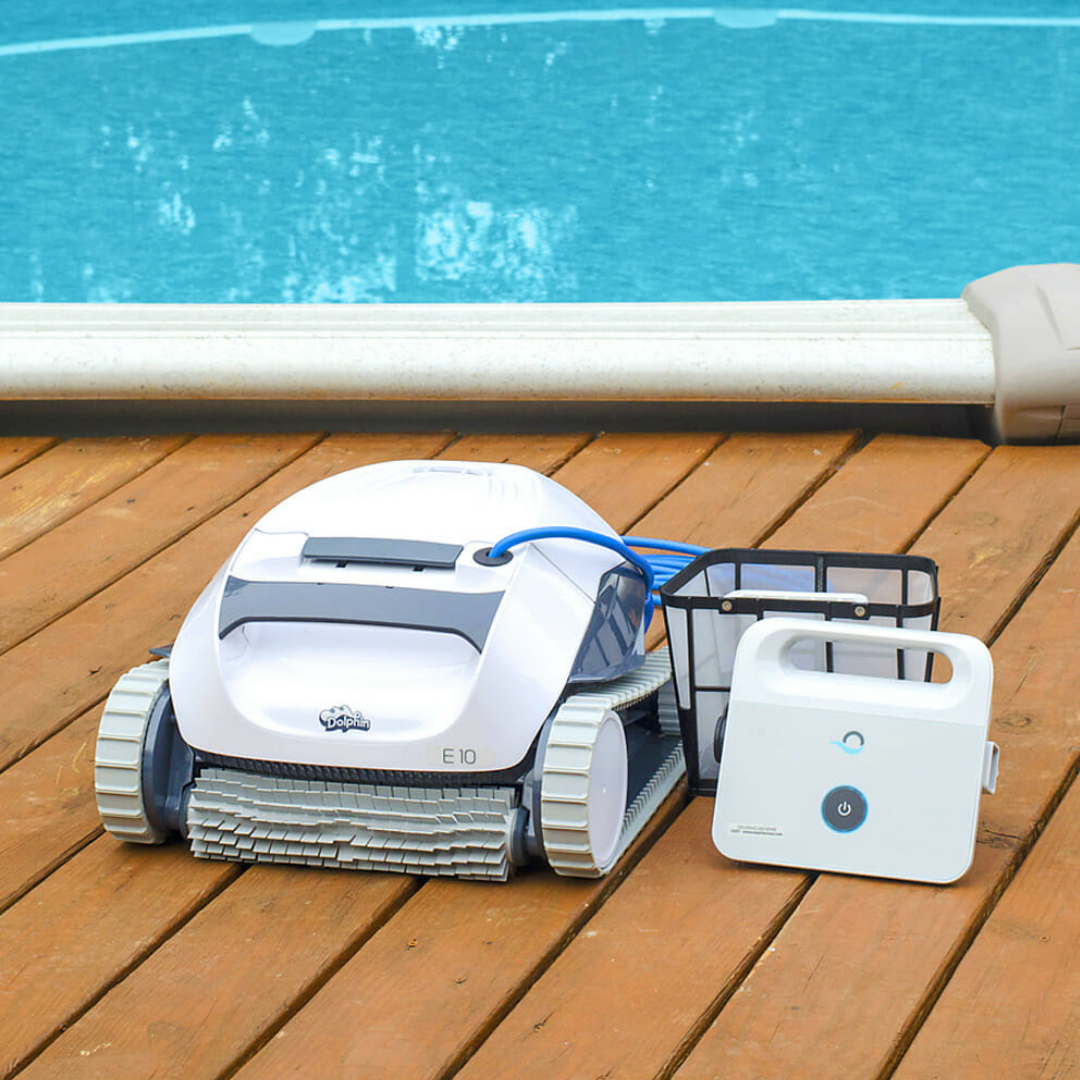Outdoor lifestyle view of the Maytronics Dolphin E 10 robotic pool cleaner resting on a wooden deck by an above-ground pool, alongside its net filter basket and power supply unit.
