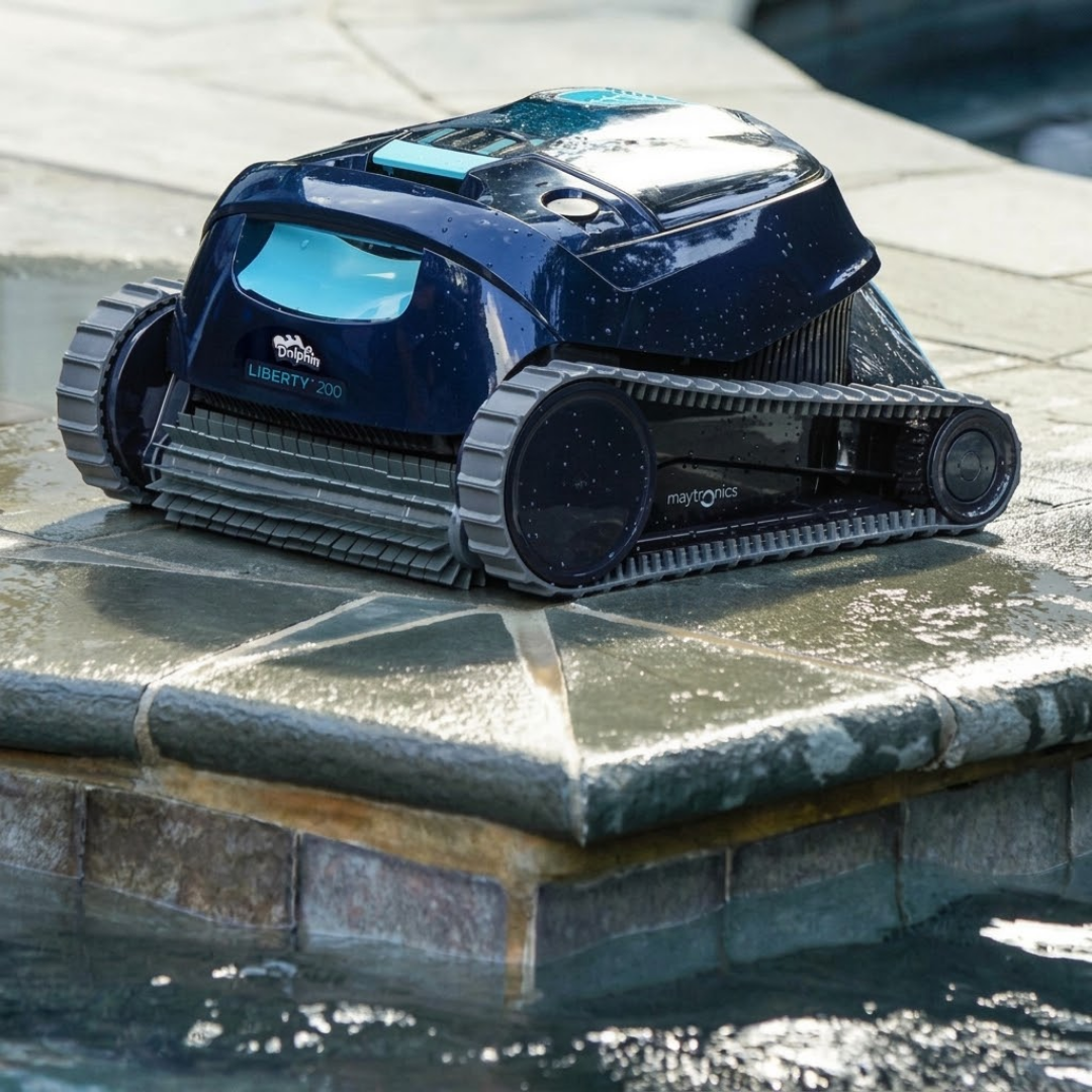 Lifestyle view of the Maytronics Dolphin Liberty 200 robotic pool cleaner resting on stone pool coping right at the water's edge, highlighting its black casing with teal accents, front scrubbing brush roll, and grey side tracks.