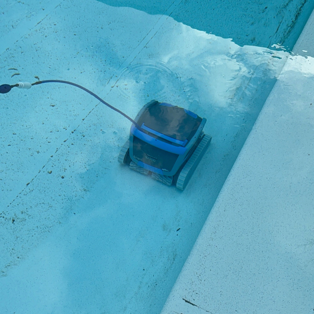 High-angle underwater shot of the dark grey Maytronics Dolphin M 700 robotic pool cleaner navigating across a light blue pool floor near the shallow end wall, showing its blue power cable trailing behind.