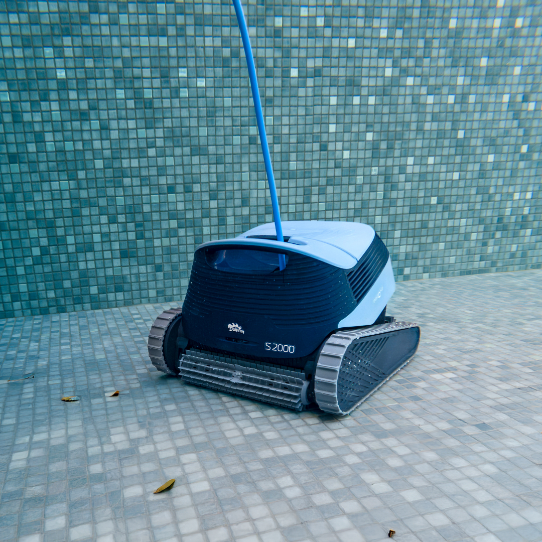 Underwater front three-quarter view of the Maytronics Dolphin S2000 robotic pool cleaner resting on a blue mosaic tiled pool floor with scattered organic debris near the pool wall, featuring its front active brush, rubber track drive system, S2000 model branding, and blue power cable rising to the surface.