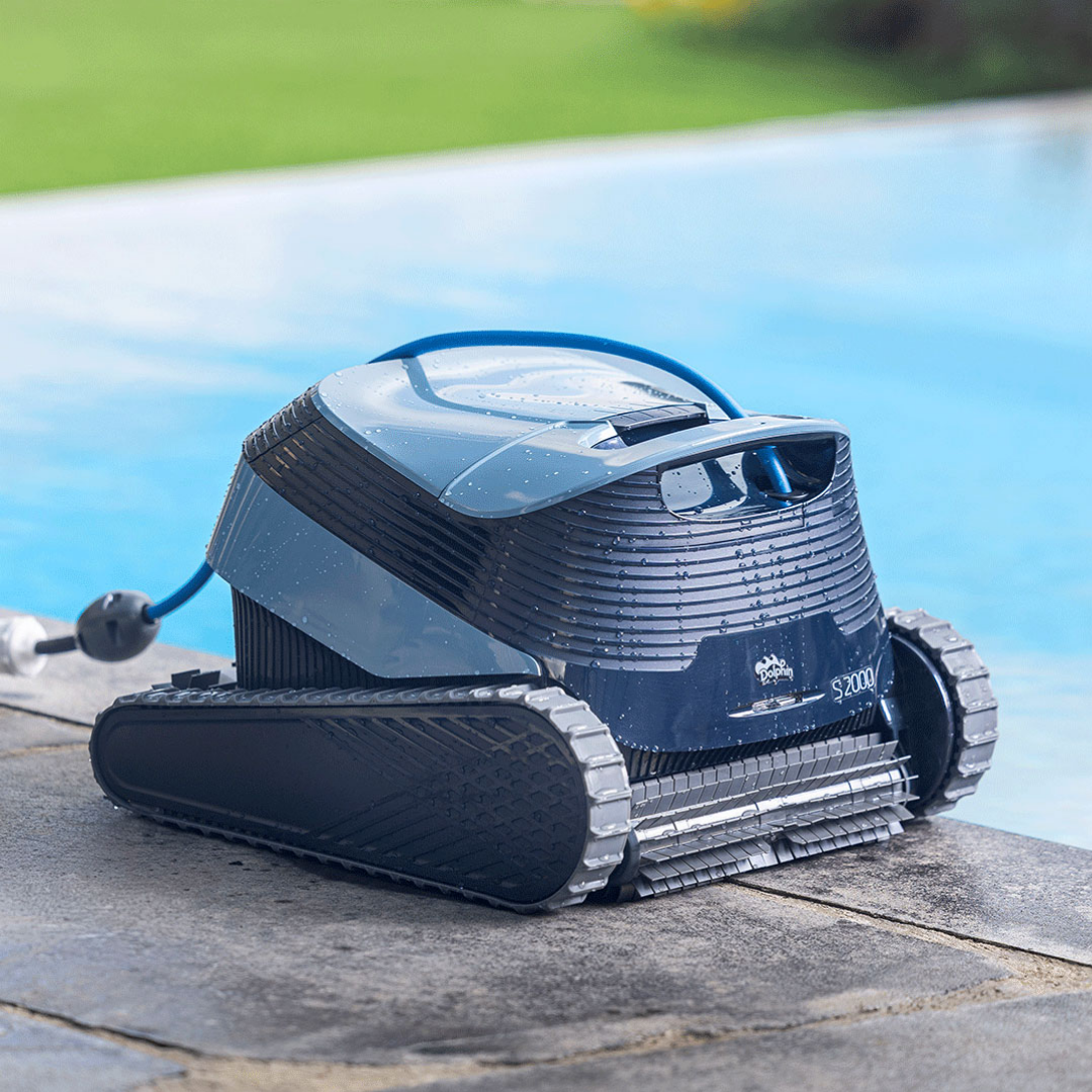 Low-angle outdoor lifestyle view of the Maytronics Dolphin S2000 robotic pool cleaner resting wet on stone coping by the pool edge, showing its front scrubbing brush, rubber continuous tracks, dark grey body with water droplets, slate blue top hatch, swivel cable, and Dolphin S2000 branding.