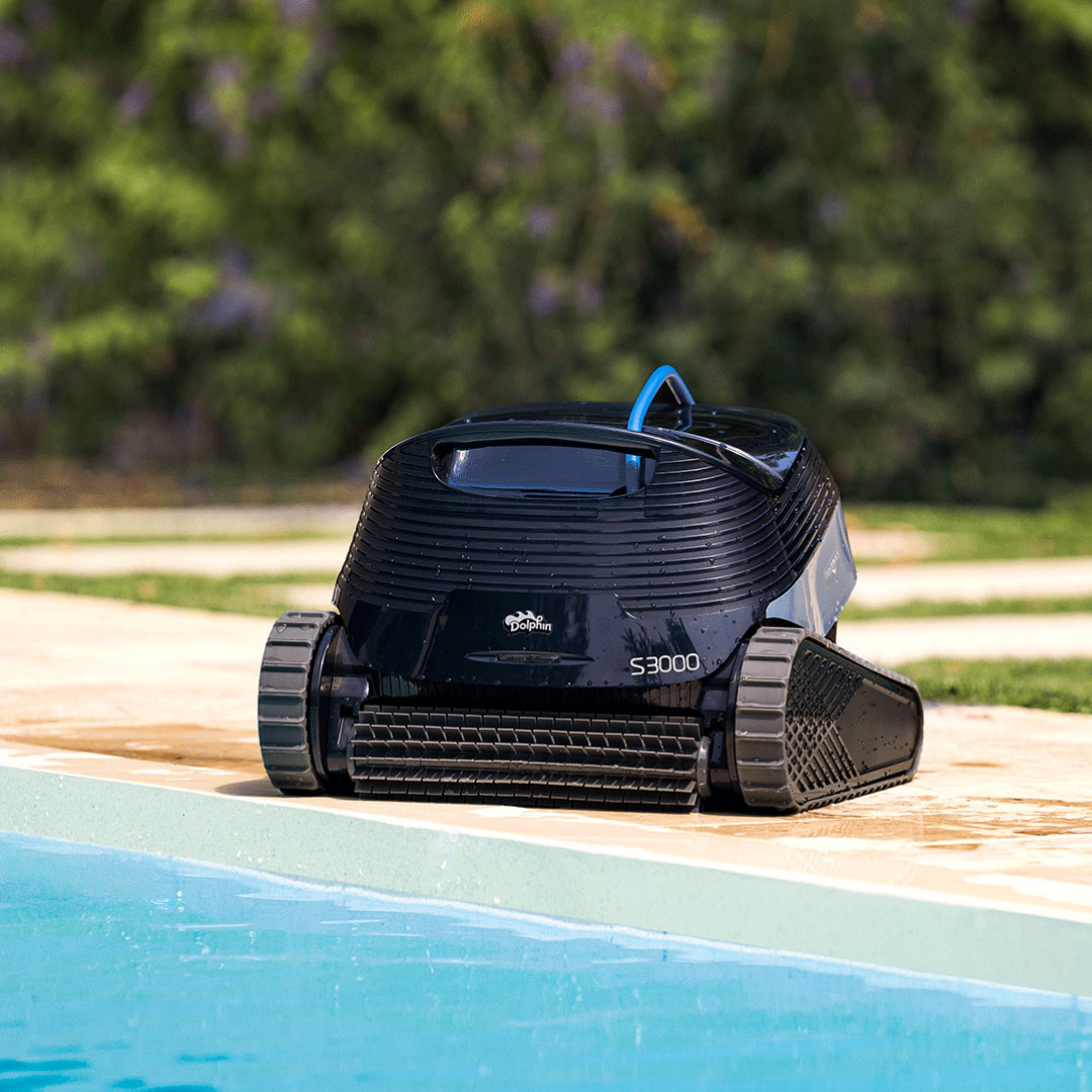 Ground-level outdoor lifestyle view of the Maytronics Dolphin S3000 robotic pool cleaner resting at the edge of a pool on stone paving, highlighting its front active scrubbing brush, dark navy chassis, Dolphin S3000 branding, rubber tracks, and blue power cable with green foliage in the background.