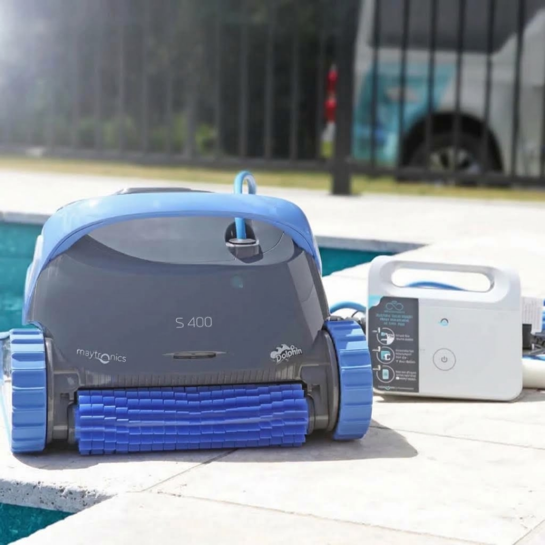 Poolside lifestyle view of the dark grey and blue Maytronics Dolphin S 400 robotic pool cleaner sitting on stone coping next to its white power supply control unit.