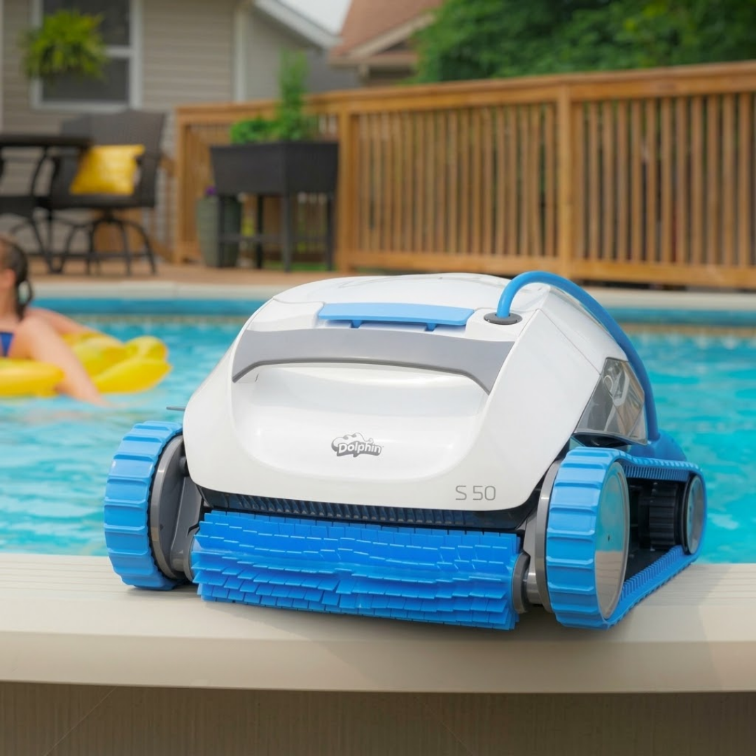 Front three-quarter lifestyle view of the white and blue Maytronics Dolphin S 50 robotic pool cleaner sitting on the edge of a backyard pool coping, with its blue active scrubbing brush and continuous tracks visible.