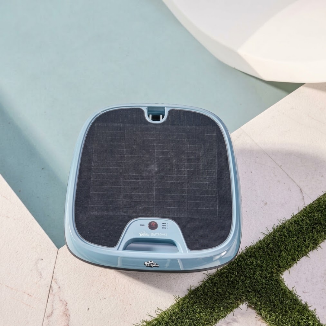 Top-down outdoor lifestyle view of the Maytronics Dolphin Skimmi 300 robotic pool skimmer resting at the pool edge on stone coping near a grass inlay, highlighting its grid-patterned solar panel top, slate blue housing, handle release latch, and front control module.