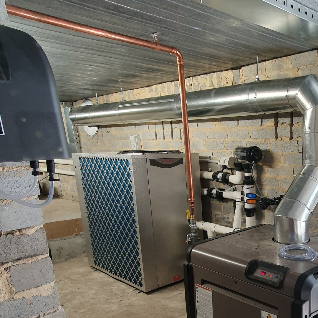 Indoor plant room installation of a metallic Rheem Thermal pool heat pump, set alongside copper pipework, ventilation ducting, PVC plumbing lines, and additional pool heating equipment against an exposed brick wall.