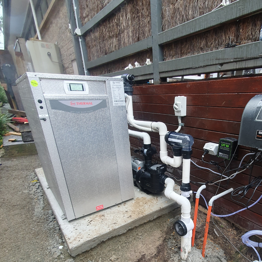 Residential outdoor installation of a Rheem Thermal pool heat pump set on a concrete slab, showcasing white PVC plumbing pipes, water circulation pump, electrical connections, and wall-mounted controllers against a wooden retaining fence.