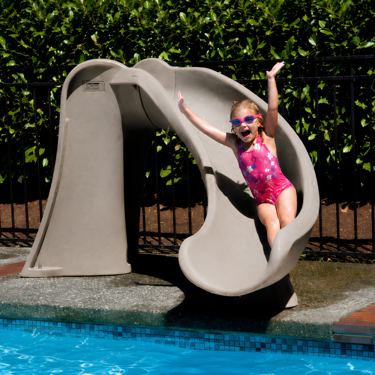 A joyful young girl wearing pink sunglasses and a floral swimsuit slides down a tan sandstone granite S.R. Smith Cyclone pool slide with her arms raised. The deck-mounted water slide curves to the right above a blue tiled inground swimming pool, with a black fence and green privacy hedge in the background.