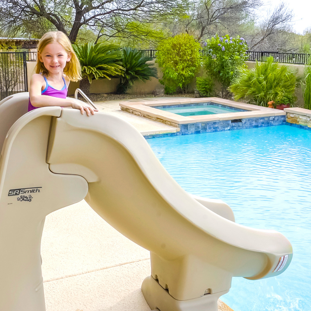 A young girl prepares to slide down a taupe S.R.Smith SlideAway removable pool slide installed next to an inground swimming pool and outdoor spa.