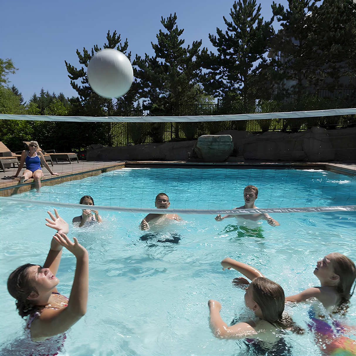 Family and friends playing with a white water volleyball over an S.R. Smith Swim N' Spike volleyball net inside a large backyard swimming pool.