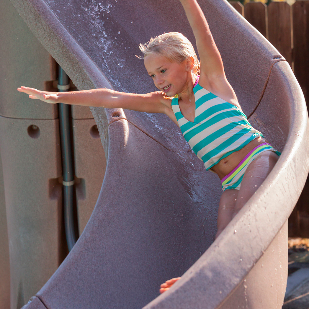 A young blonde girl wearing a striped teal and white swimsuit riding down an S.R. Smith TurboTwister sandstone tan residential swimming pool slide with active running water, arms outstretched with a wooden fence in the background.