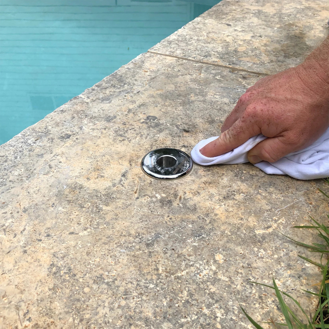 Close-up view of a hand using a white cloth to wipe down a chrome flush-mount deck sleeve bracket installed into the stone coping edge of an inground swimming pool.
