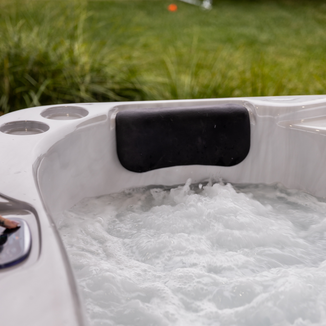 Tropic Seas Spas Lounge series charcoal gray headrest pillow installed in a white acrylic hot tub with bubbling water, featuring a grassy backyard lawn backdrop.