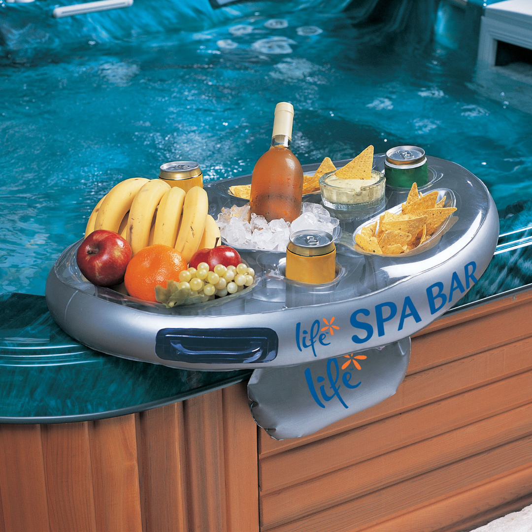 A lifestyle view of an inflatable silver Life Spa Bar snack tray and drink caddy resting on the edge of a bubbling blue acrylic hot tub. The bar is filled with a wine bottle on ice, canned drinks, tortilla chips, and fresh fruit, with its weighted stability pouch hanging over the side of the wooden spa cabinet.