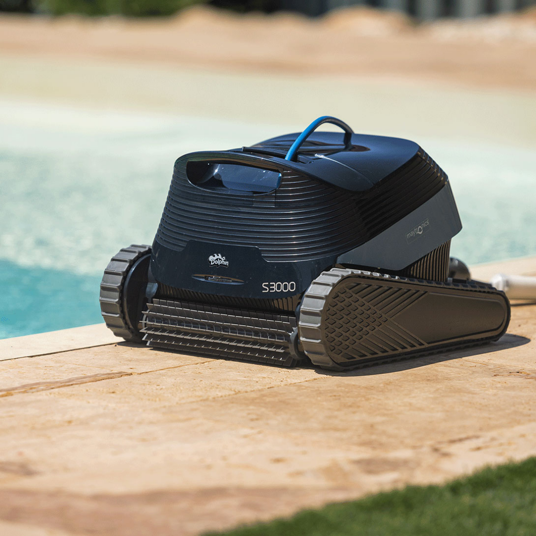 Low-angle outdoor lifestyle view of the Maytronics Dolphin S3000 robotic pool cleaner resting on stone poolside paving near the water's edge, highlighting its dark navy chassis, front active scrubbing brush, rubber continuous tracks, S3000 model branding, and blue top handle.