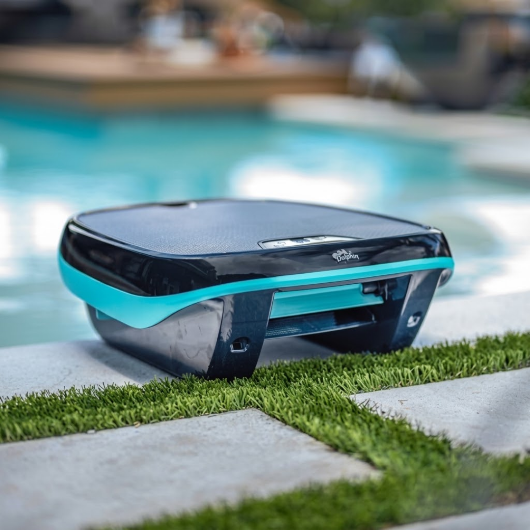 Outdoor lifestyle shot of the Maytronics Dolphin Skimmi 200 robotic pool skimmer resting poolside on a paver patio with grass inlay, showing its dark grey body, bright teal accent band, front intake flap, and solar panel top.