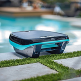 Outdoor lifestyle shot of the Maytronics Dolphin Skimmi 200 robotic pool skimmer resting poolside on a paver patio with grass inlay, showing its dark grey body, bright teal accent band, front intake flap, and solar panel top.