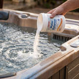 A person's hand pouring Poppits Water Balance powder directly into the bubbling water of an outdoor hot tub. The label on the 1kg container shows it is Step 2, used to raise pH and Total Alkalinity with 99-100% Sodium Bicarbonate.