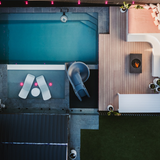 Aerial drone view of a luxury modern backyard featuring an inground swimming pool, integrated tanning ledge with chaise lounges, a grey spiral pool slide, and an adjacent timber deck lounge with a fire pit.