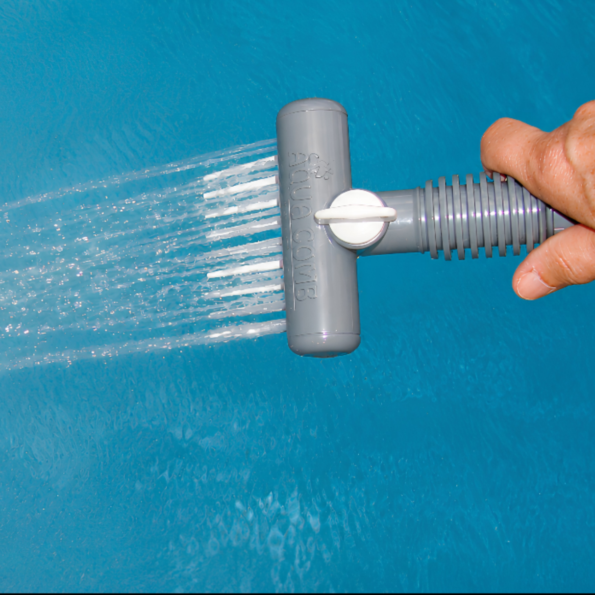 A close-up view of a hand holding a gray Aqua Comb tool as it sprays powerful parallel streams of water from its white tines over bright blue pool or hot tub water, showing the embossed Aqua Comb brand name on the sprayer head.