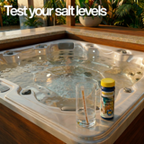 An AquaChek salt test strips bottle and a water testing glass sitting on the edge of a backyard hot tub spa, with text overlay that reads "Test your salt levels."