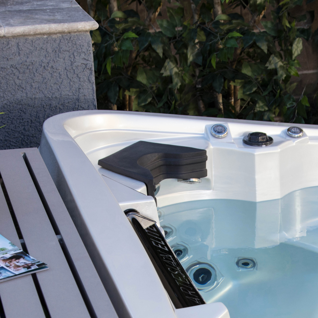 High-angle view of a charcoal black Artesian Elite neck headrest pillow installed in a white acrylic hot tub corner seat, filled with clean water next to a spa steps panel and backyard greenery.