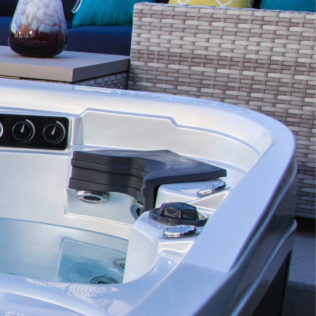 A charcoal black Artesian Elite neck headrest pillow installed in the corner seat of a luxury white acrylic hot tub, positioned over the integrated spa jets with outdoor patio furniture in the background.